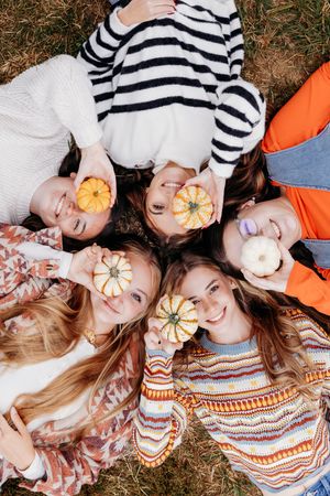 Five young women lying in a circle on grass, each holding small decorative pumpkins up to their faces.