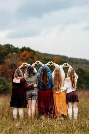Five girls stand in a field making heart shapes with their hands against an autumn landscape.