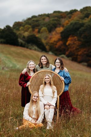 Five young women pose in a fall field with a wicker chair surrounded by colorful autumn foliage.