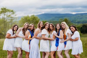 Nine smiling young women in white dresses with colorful graduation sashes pose in a green field.