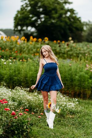 Blonde girl in blue denim mini dress with white boots walking through sunflower garden.