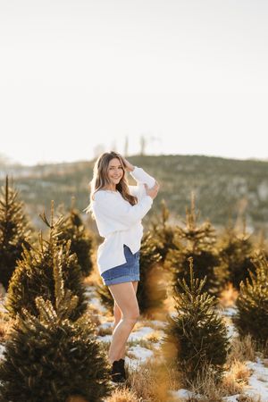 Smiling brunette in white sweater and denim skirt posing at golden-lit Christmas tree farm.