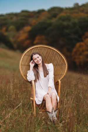 Smiling young woman in white dress sits in a wicker peacock chair in a sunlit autumn field.