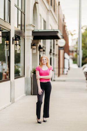 Young woman in pink top and black pants posing confidently on urban sidewalk outside brick building.