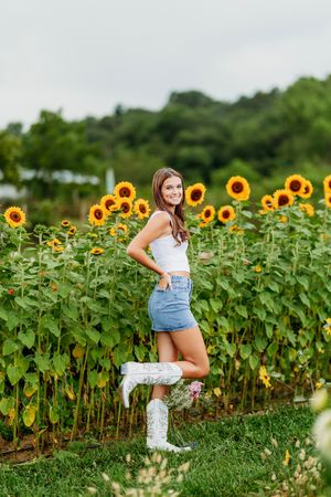 Young woman in white top, denim skirt, and white cowboy boots posing in a sunflower field.