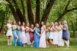 Large group of women in colorful spring dresses pose together under a large tree outdoors.