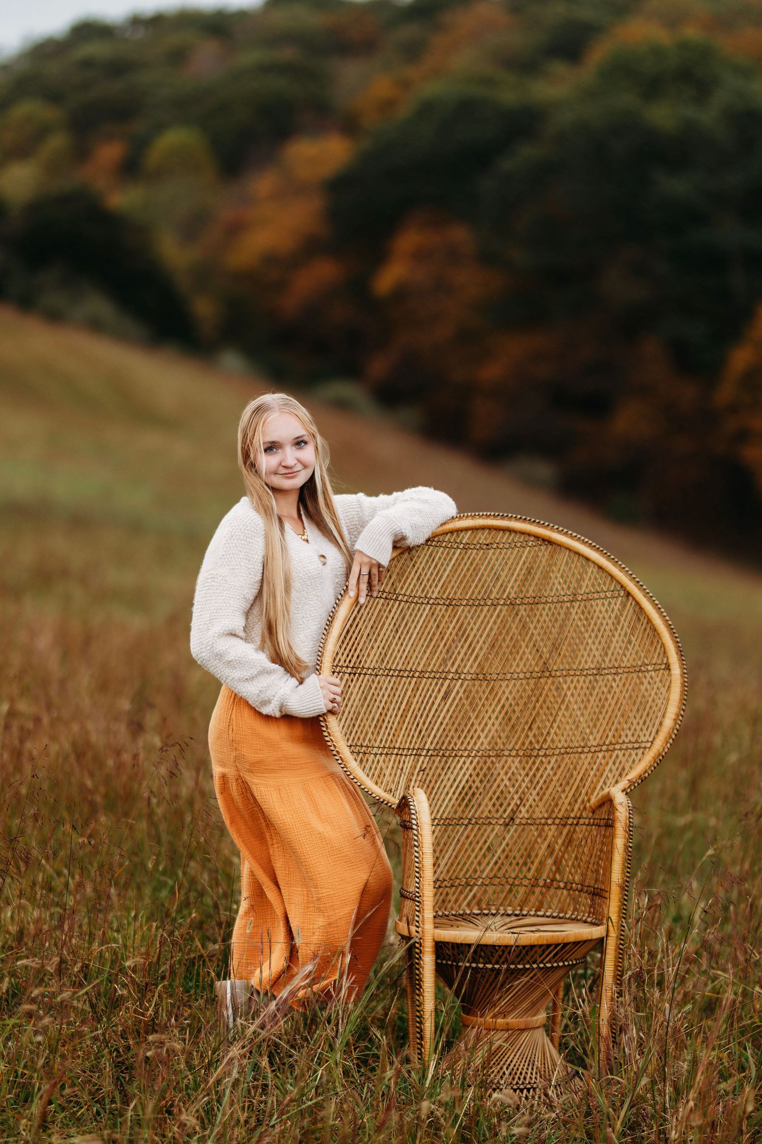 Blonde teen in white sweater and orange skirt leans on a wicker peacock chair in an autumn field.