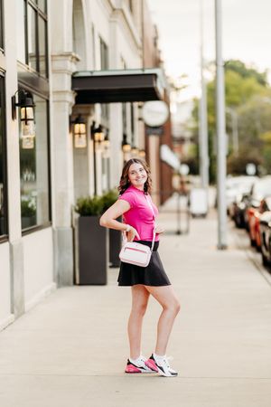 Smiling teen in pink polo and black skirt holding pink bag on city sidewalk.