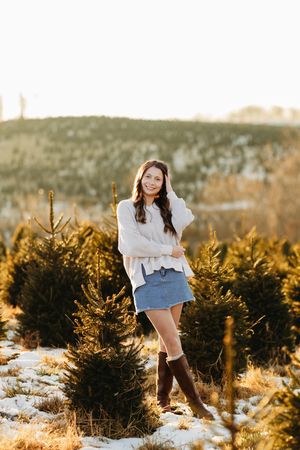 Brunette woman in white sweater and denim skirt posing at snowy Christmas tree farm.