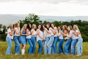 Group of women in white tops and jeans laugh together in a field with mountain views behind them.