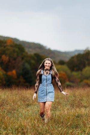 Smiling young woman in denim pinafore and lace top walks through an open field with autumn foliage.