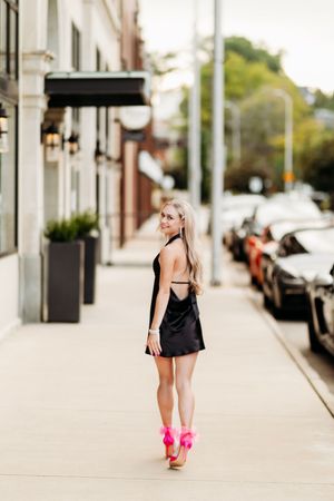 Blonde woman in black backless mini dress with pink heels posing on urban street.