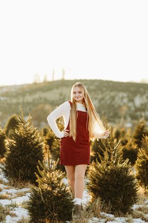 Blonde teen girl in red dress posing among snow-dusted Christmas trees at sunset.