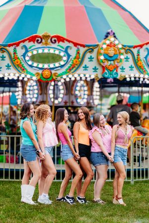 Six teenage girls in pink tops and denim shorts laugh together in front of a colorful carousel.