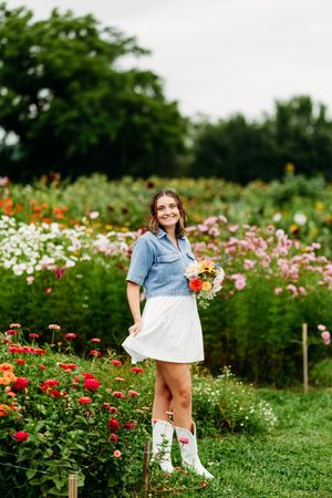 Smiling young woman in blue shirt and white skirt holding bouquet in a vibrant flower field.