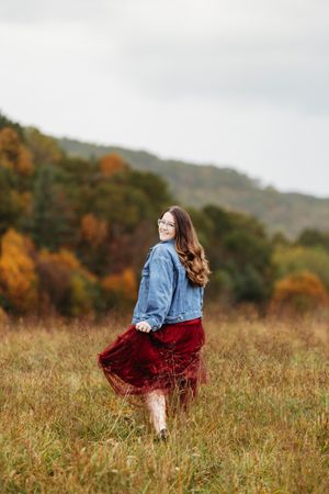 Young woman in denim jacket and burgundy skirt twirling in autumn field with colorful fall foliage.