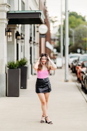 Young woman in pink bodysuit and black leather skirt posing on city sidewalk.
