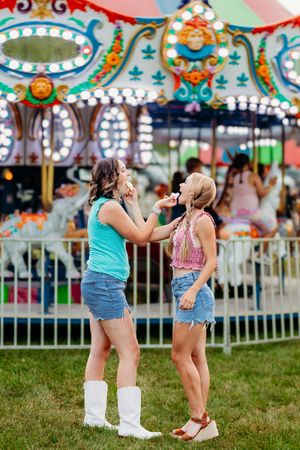 Two young women share cotton candy in front of a brightly lit carousel at a summer fair.