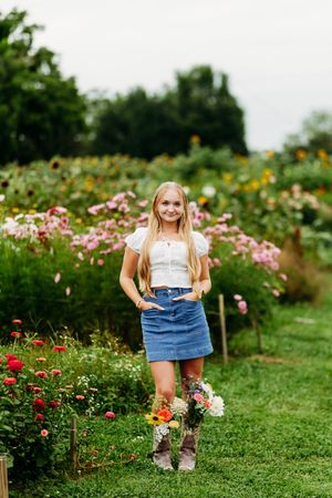 Smiling blonde girl in white top and denim skirt holding flowers in a colorful flower garden.