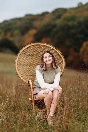 Smiling young woman sitting in wicker peacock chair in golden autumn meadow with fall trees.