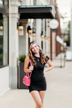 Smiling brunette in black mini dress carrying pink handbag on urban sidewalk.