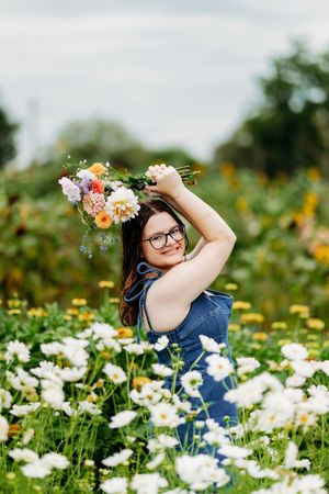 Young woman with glasses holding a colorful bouquet overhead while standing in a white flower field.