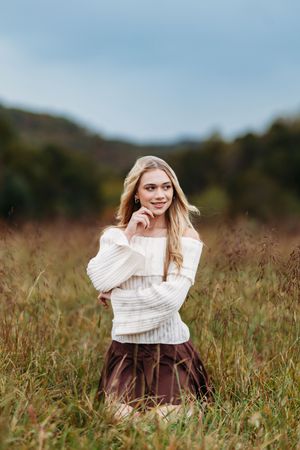 Blonde woman in cream off-shoulder sweater and brown skirt posing in tall grass autumn field.