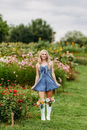 Smiling blonde woman in denim dress and white cowboy boots holding flowers in a colorful garden.