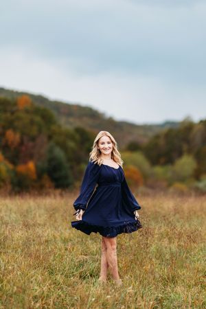 Smiling blonde woman twirls in a navy blue dress in an open field with autumn foliage backdrop.