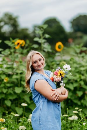 Smiling blonde woman in blue dress cradling a colorful bouquet in a sunflower garden.