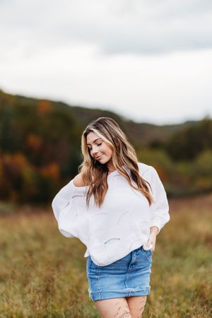 Young woman in white oversized sweater and denim skirt poses in an autumn field with hills behind.