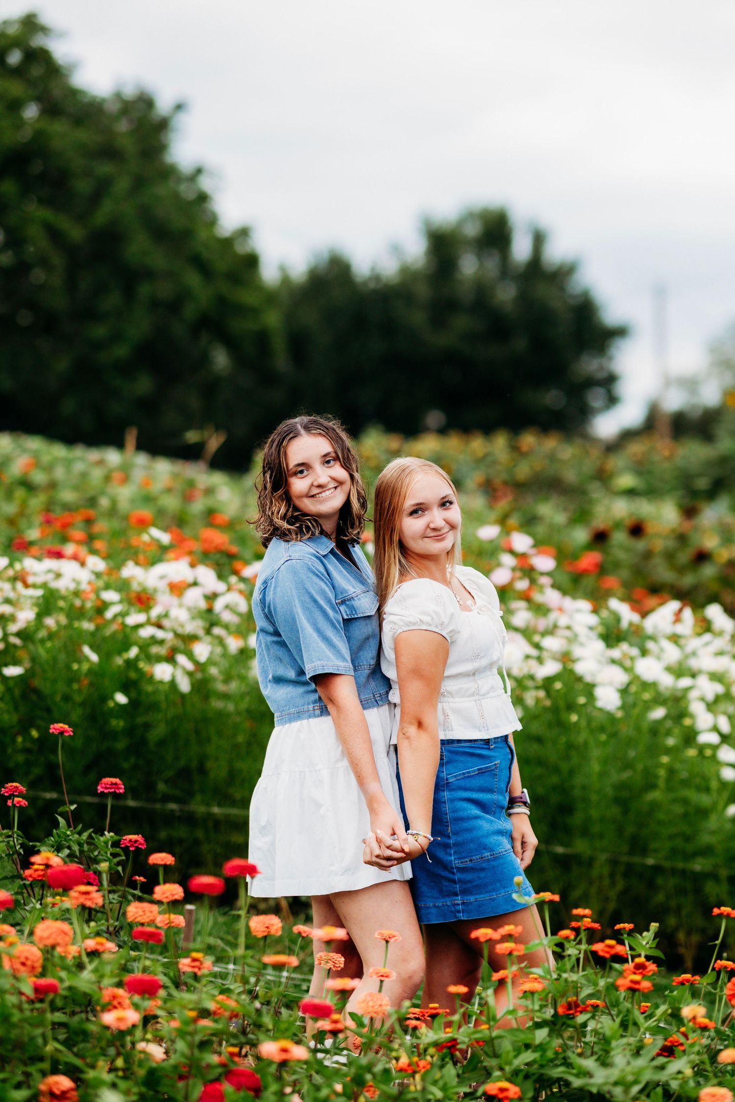 Two young women holding hands in a colorful flower field wearing denim and white outfits, smiling at camera.