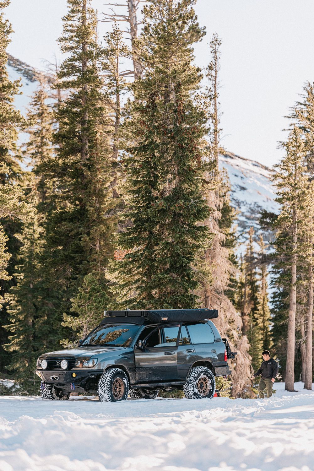 Black SUV parked in snowy mountain terrain surrounded by tall pine trees and snow-covered slopes at sunset.