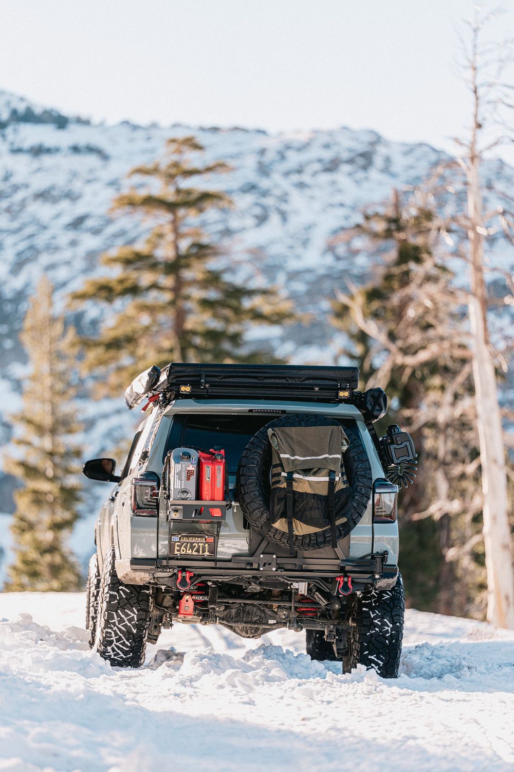 Off-road vehicle with roof rack and gear navigating through snowy mountain terrain with evergreen trees in background.