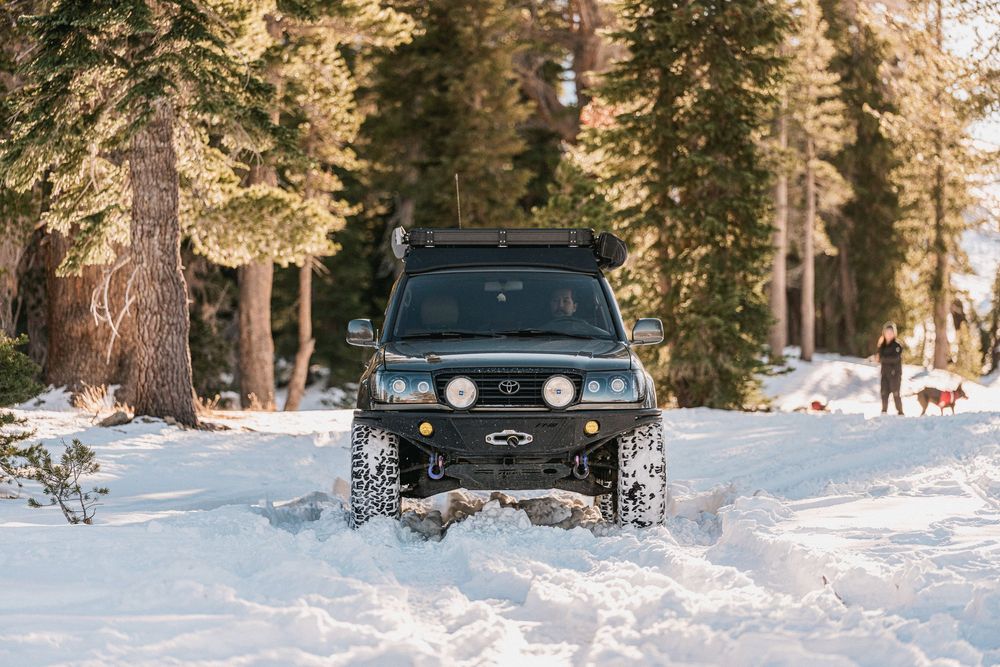 Black SUV driving through deep snow on a forest road during winter with tall evergreen trees in the background.