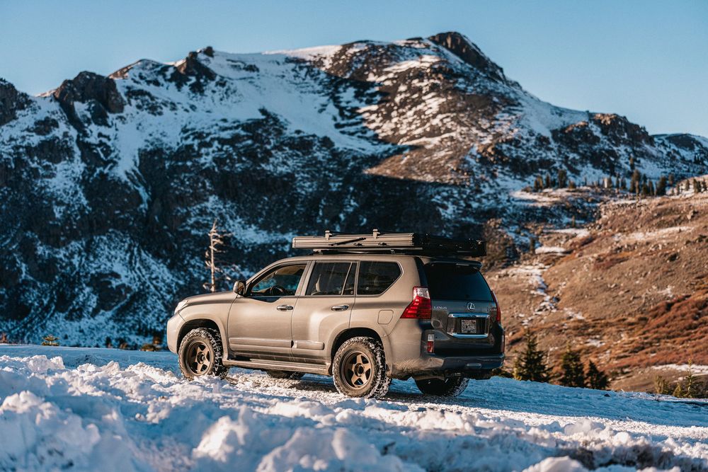 A silver SUV parked on a snowy mountain road with rugged snow-capped peaks in the background at sunset.