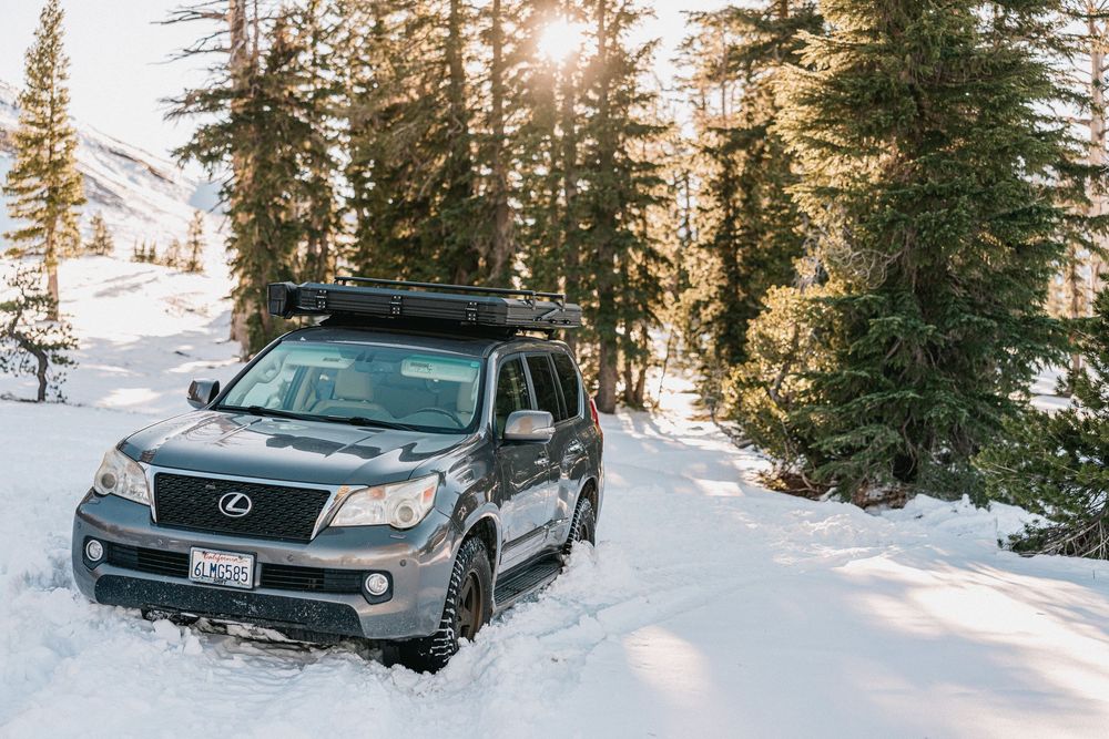 A dark green SUV parked in a snowy mountain forest with sunlight streaming through evergreen trees.