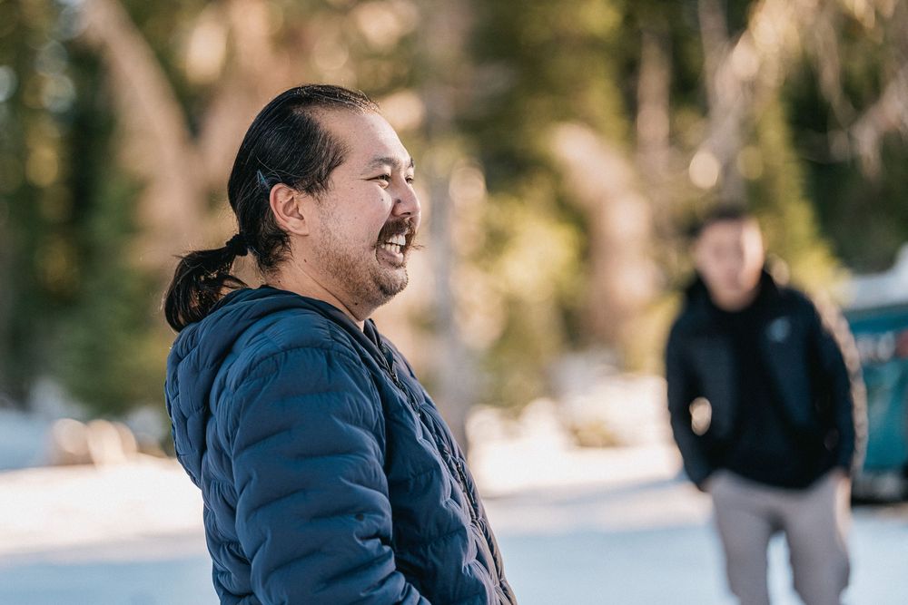 A person in a blue winter jacket laughs joyfully outdoors in a snowy setting with someone visible in the background.