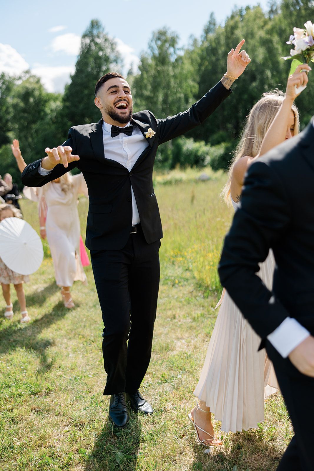 Joyful wedding party celebrates in a sunny meadow with energetic dancing and raised arms.