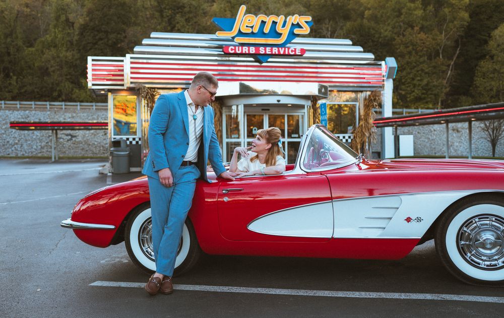 A vintage red and white classic car parked at a retro drive-in restaurant with neon signage at dusk.
