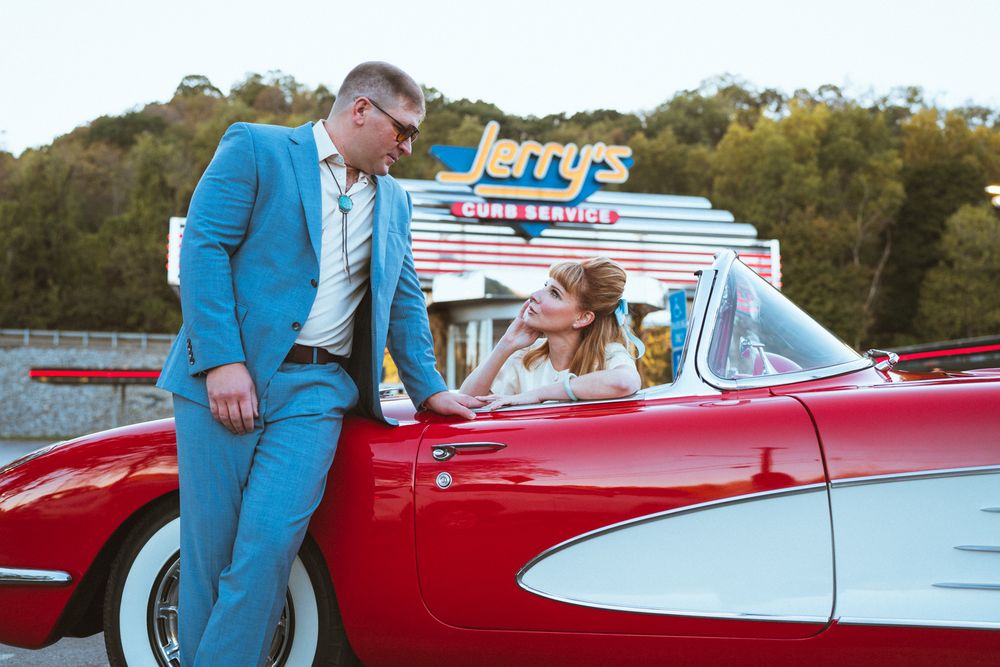 Vintage red and white classic car parked outdoors with people in bright blue attire gathered around it.