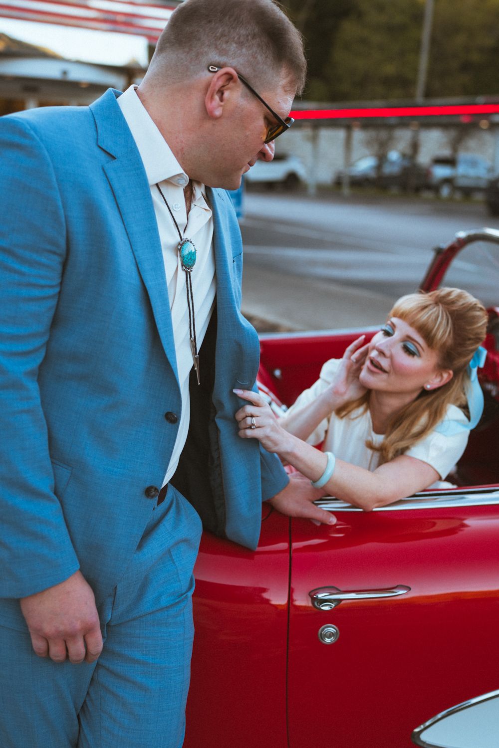 A couple in vintage style clothing share a romantic moment next to a classic red convertible car.