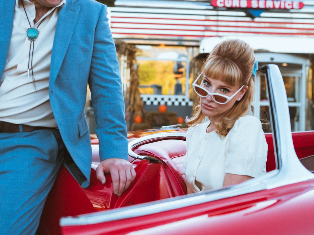 A person in a blue suit helps someone into a vintage red convertible car outside a diner.