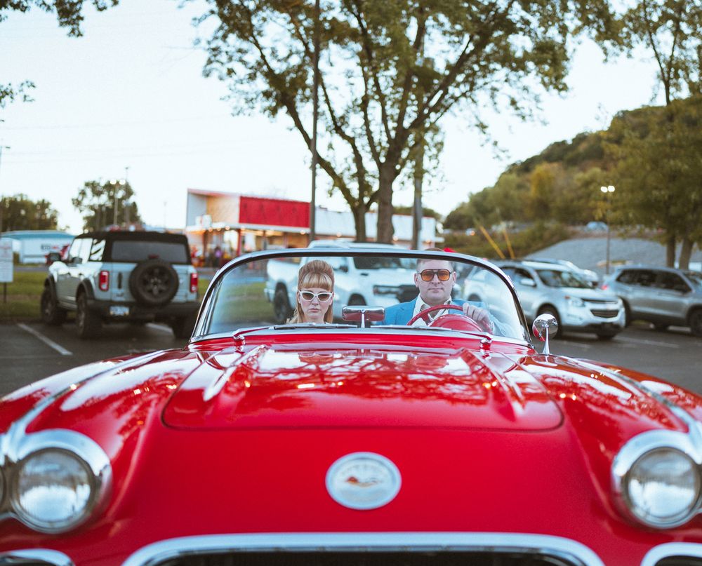 A shiny red vintage automobile with chrome accents photographed under trees in a parking area.