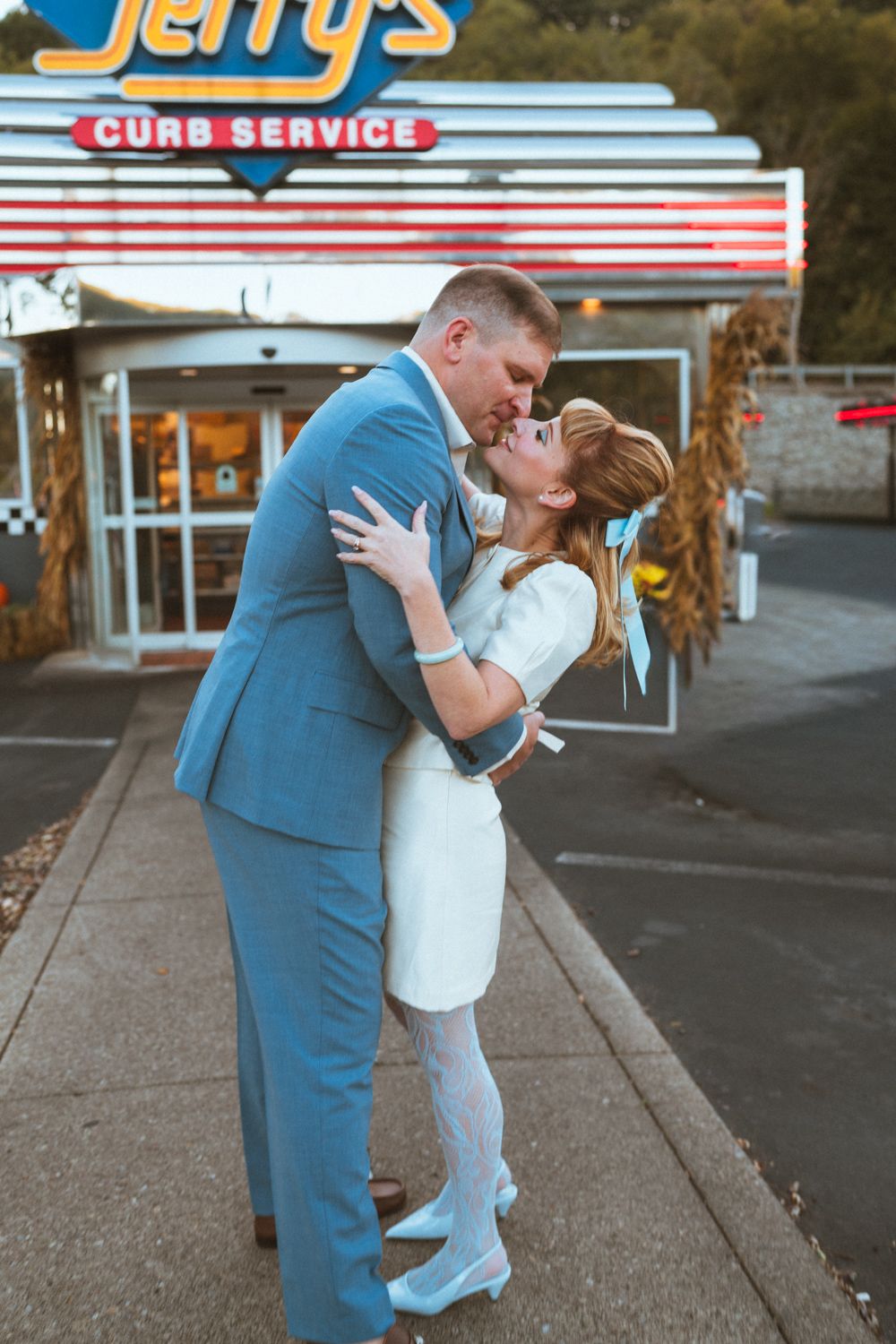 A romantic couple embraces and kisses in front of a vintage drive-in restaurant with retro signage and curb service.