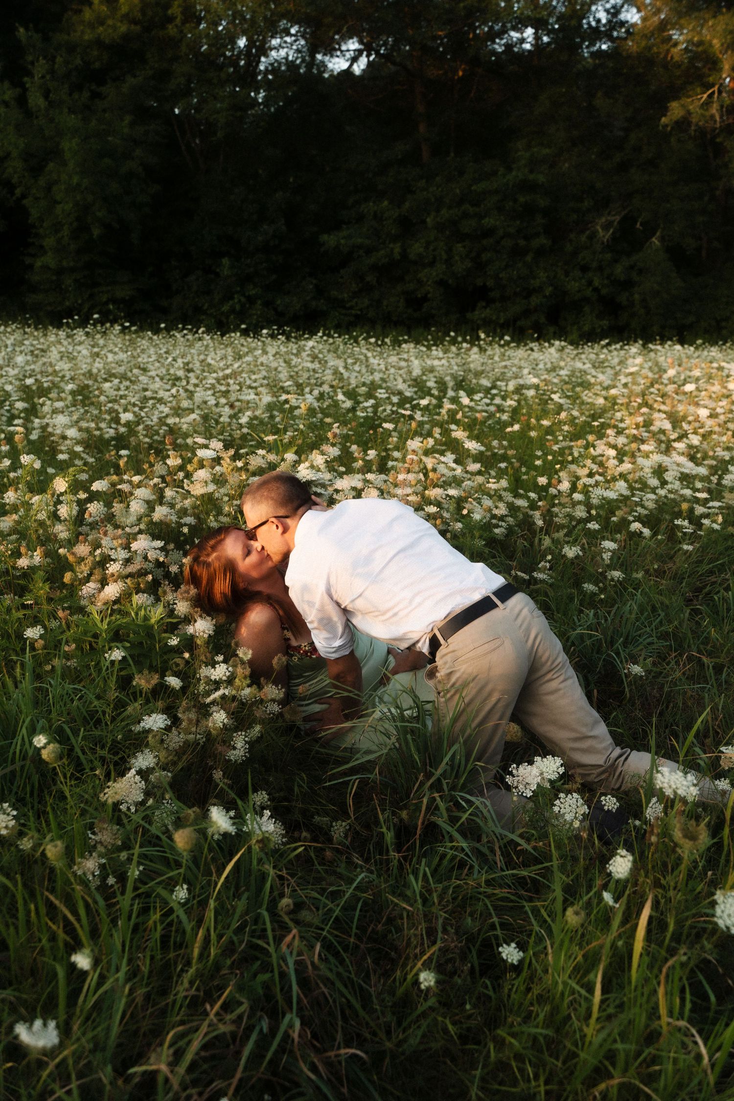 Two people share an intimate moment lying in a field of white wildflowers at sunset.
