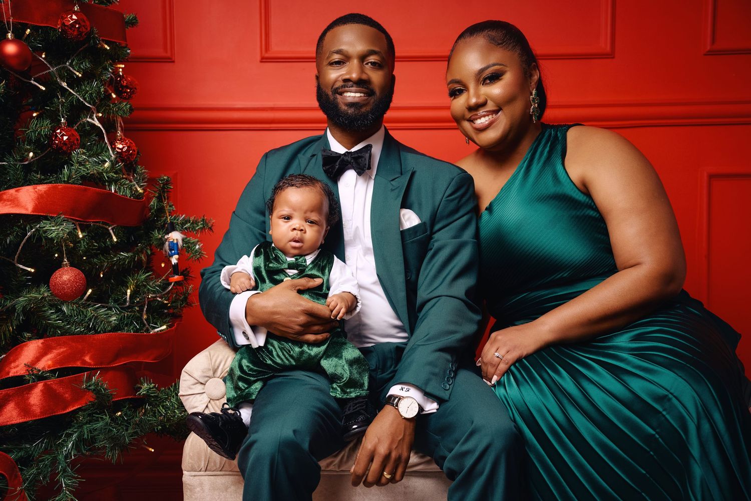 A couple in elegant emerald green attire poses for romantic holiday photos in front of a red wall and Christmas tree.