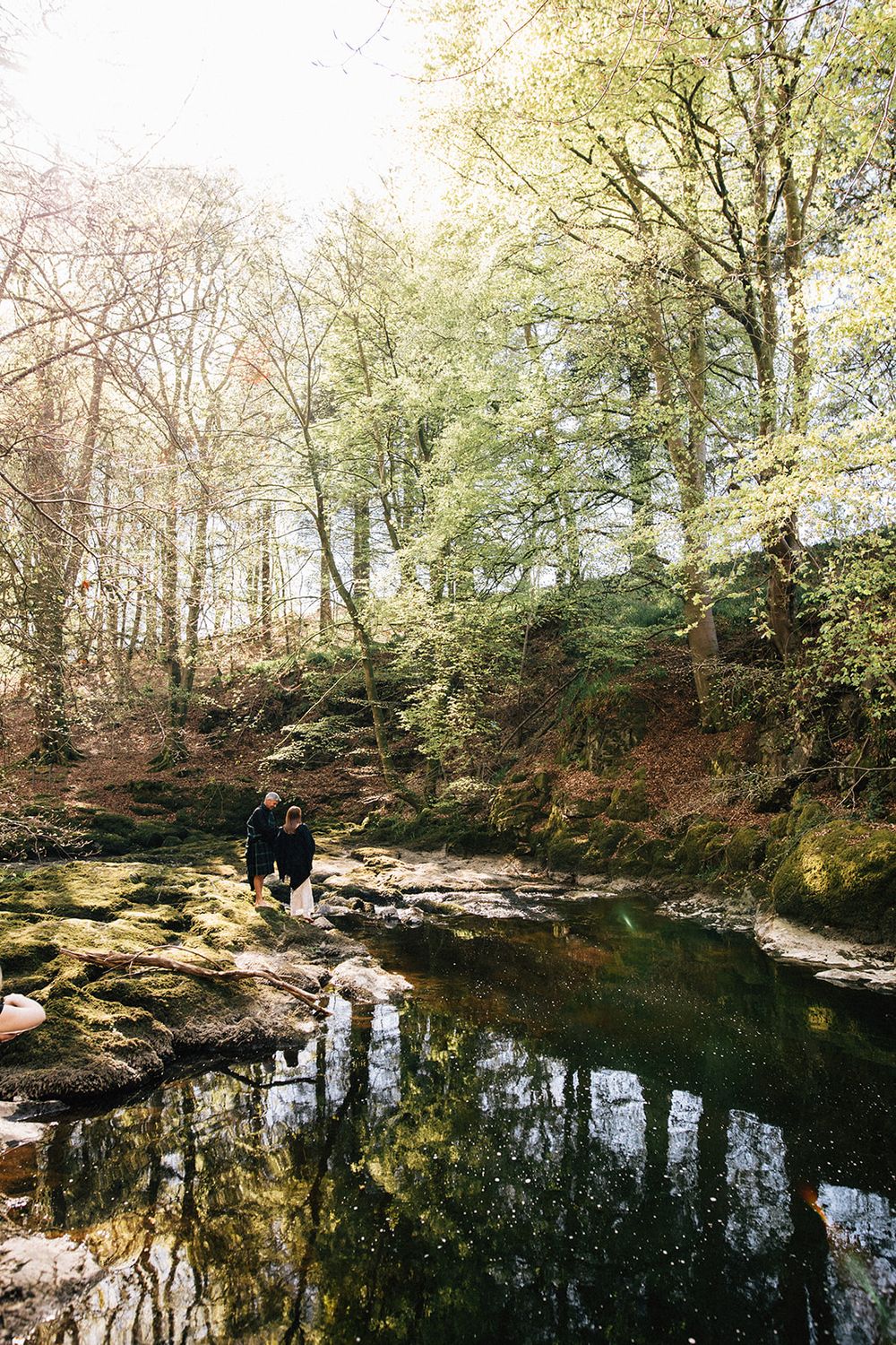 Two people stand beside a tranquil forest pool reflecting green trees and moss-covered rocks in dappled sunlight.