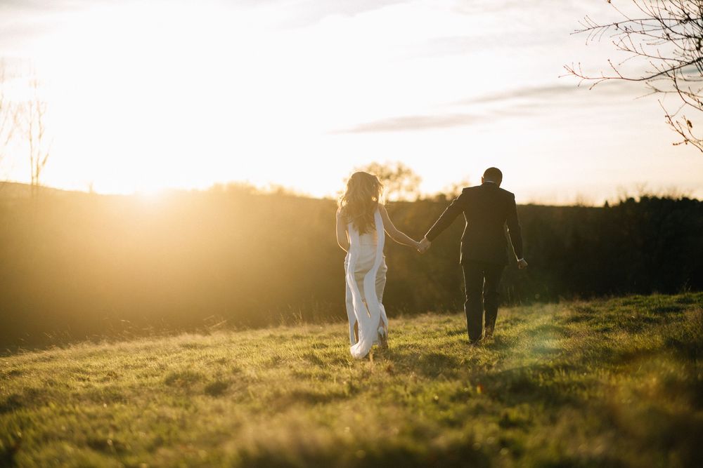 Couple holding hands walking through golden sunset field, bride in white dress, romantic outdoor wedding photo.
