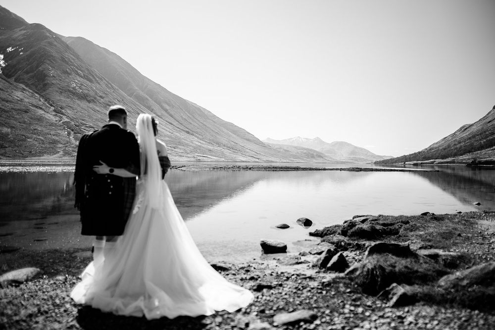 A couple in wedding attire stands silhouetted against a misty Scottish loch and mountains.